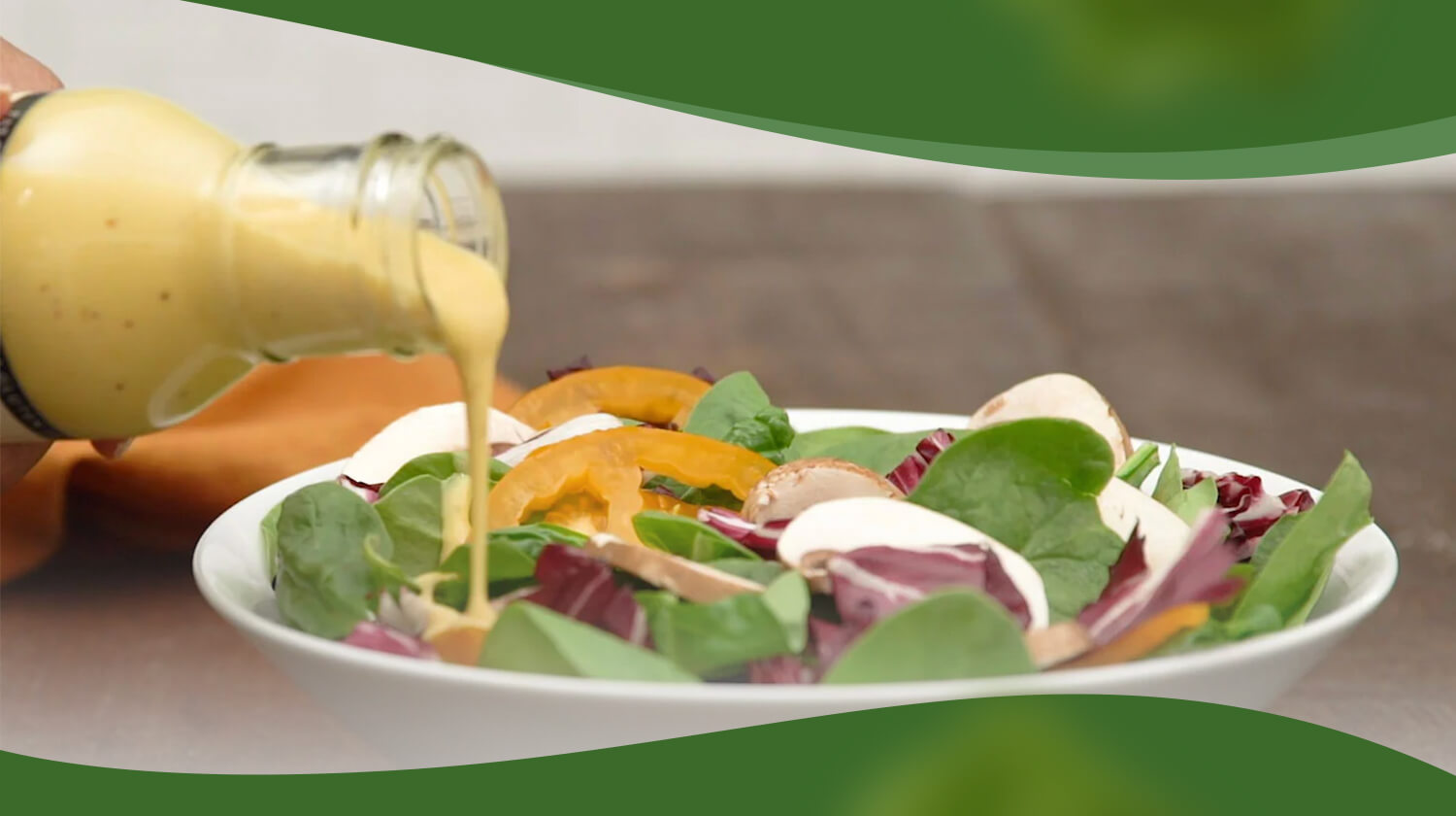 A bottle of golden dressing is being poured onto a fresh salad with spinach, radicchio, tomatoes, and mushrooms, set against a wooden table.