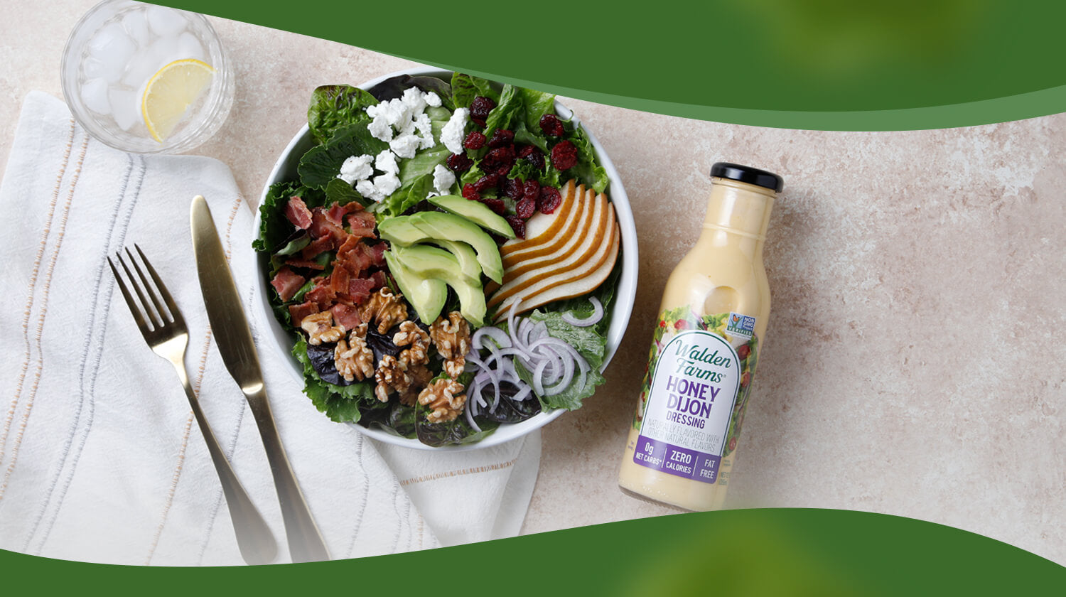 A colorful salad with avocado, pear slices, walnuts, feta, and cranberries is served on a bowl next to a fork and knife, with honey Dijon dressing nearby.
