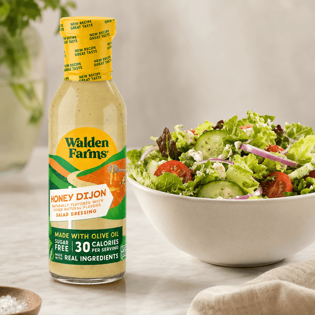 Walden Farms Honey Dijon Salad Dressing made with olive oil displayed beside a fresh salad with cucumbers, tomatoes, onions, and crumbled cheese.