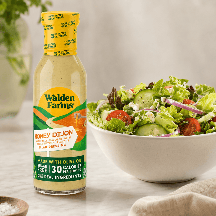Walden Farms Honey Dijon Salad Dressing made with olive oil displayed beside a fresh salad with cucumbers, tomatoes, onions, and crumbled cheese.