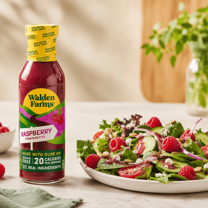 Walden Farms Raspberry Vinaigrette made with olive oil beside a mixed green salad topped with raspberries, cucumbers, tomatoes, onions, feta cheese, and vinaigrette.